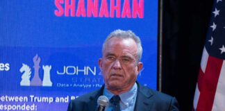 A man speaking into a microphone at an event with a large screen behind him displaying data