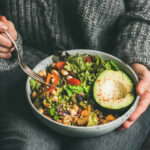 A person holding a bowl of colorful salad with avocado and greens