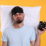 The Surprising Sleep Duration for Optimal Health Man wearing a sleep mask holding an alarm clock with a frustrated expression