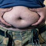 Metabolism Mystery Solved: Hormone Ignites Fat Loss Man holding his belly with both hands, wearing a blue shirt and camouflage pants