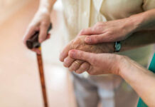 Two hands clasped together, one elderly and one younger, with a cane in the background
