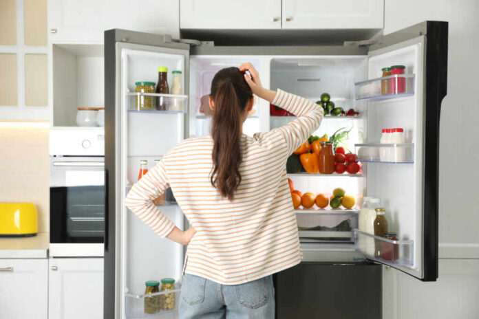 Young,Woman,Near,Open,Refrigerator,In,Kitchen,,Back,View A woman standing in front of an open refrigerator filled with various fruits and vegetables