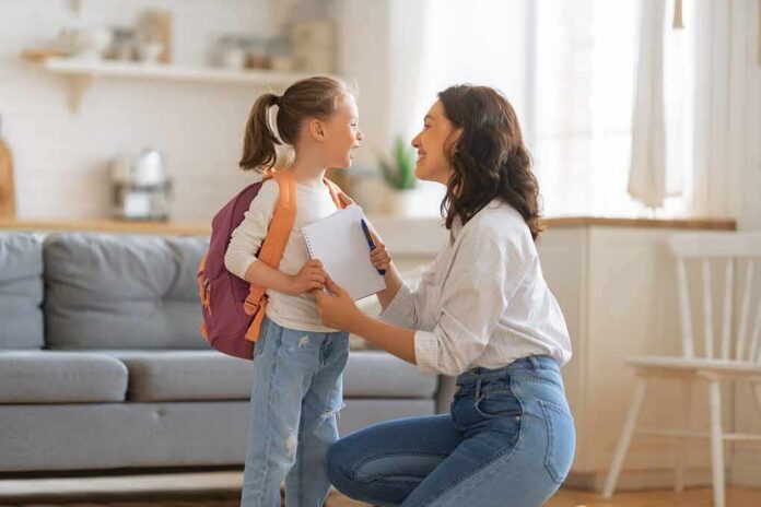 shutterstock_2026477766-1 Mother kneels to talk with daughter wearing backpack