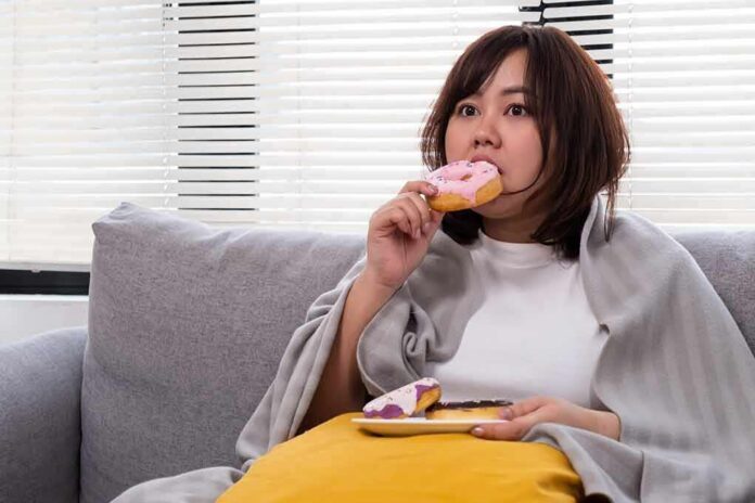 shutterstock_2158040957 Woman on couch eating a pink frosted donut